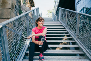 Woman in workout gear sitting on outdoors stairs holding a water bottle. Weight loss before a tummy tuck.