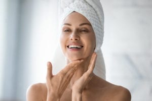 Tips to Plan a Facelift. Happy woman with her hair wrapped in a towel looking at her face in the bathroom mirror.