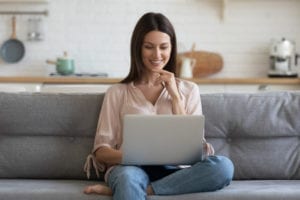 Smiling young woman sitting on her couch at home using laptop to research how to pick a plastic surgeon.